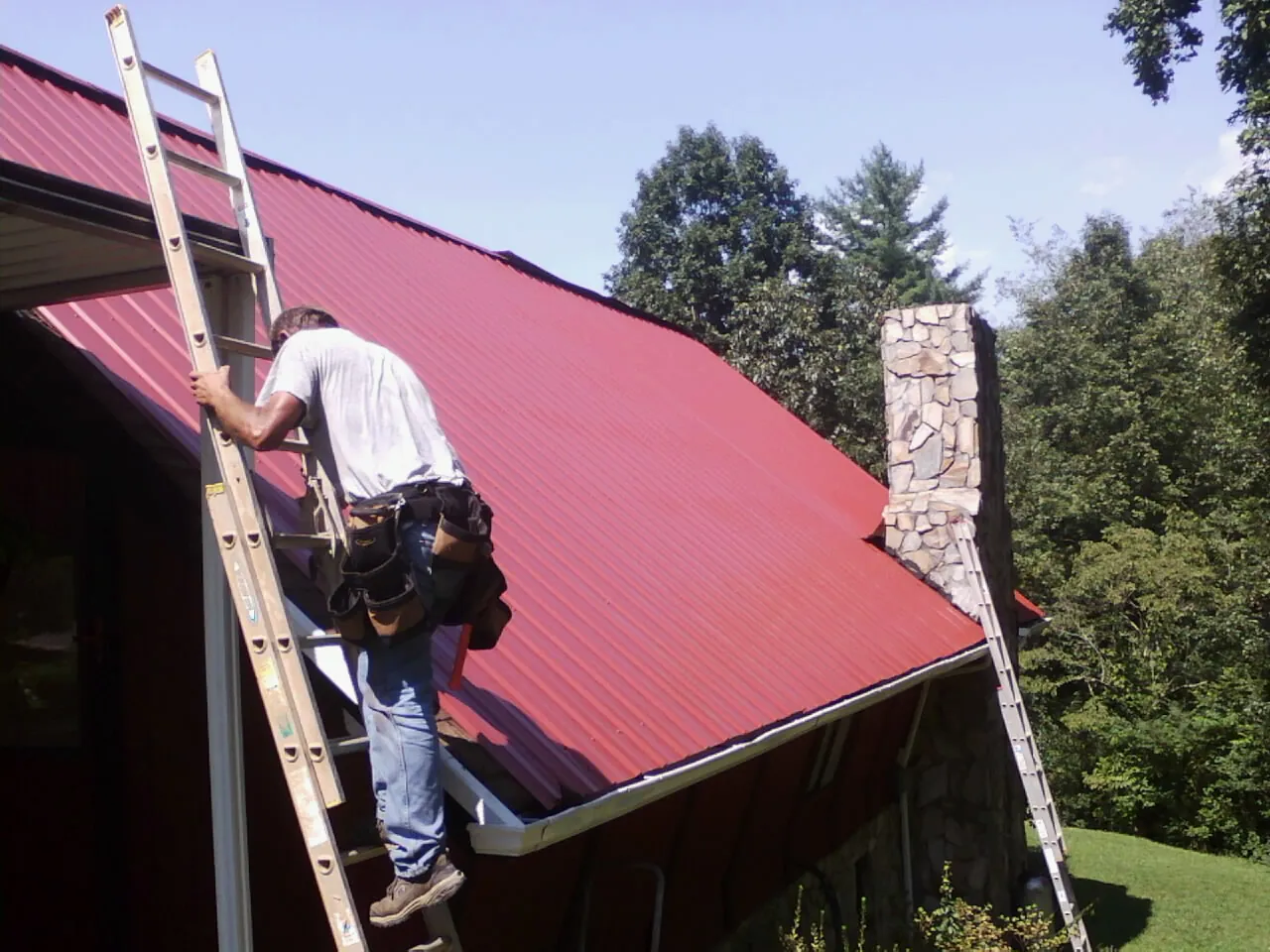 Roofer on a ladder beside a red metal roof and stone chimney — Soffit & Fascia Repair by Roofing Brothers in Coupland